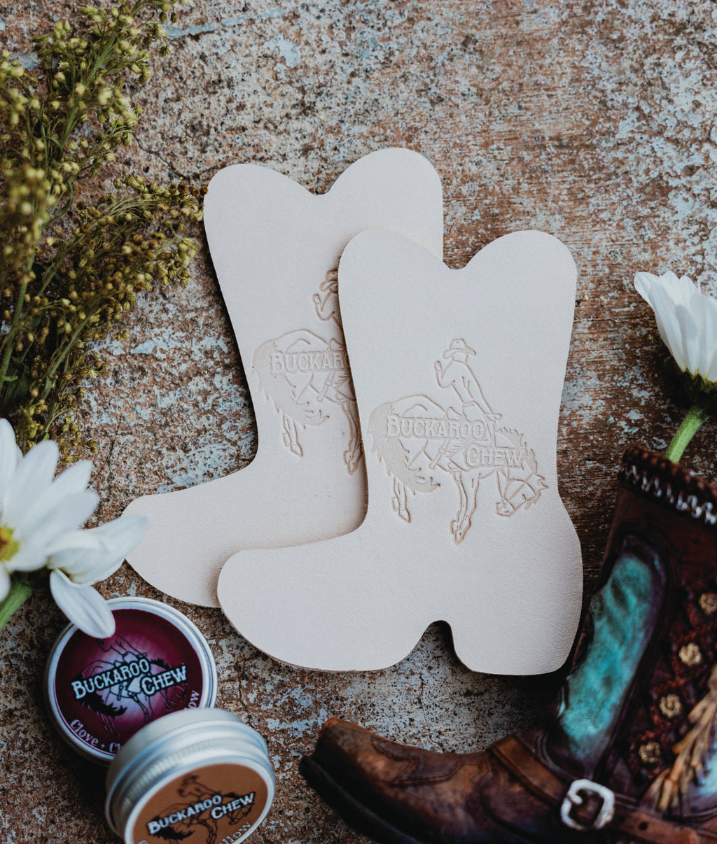 Two beige leather cowboy boot-shaped items with embossed Buckaroo Crew graphics, arranged on a rustic textured surface with a brown leather boot, white flowers, and small tins of Buckaroo Crew lip balm nearby.