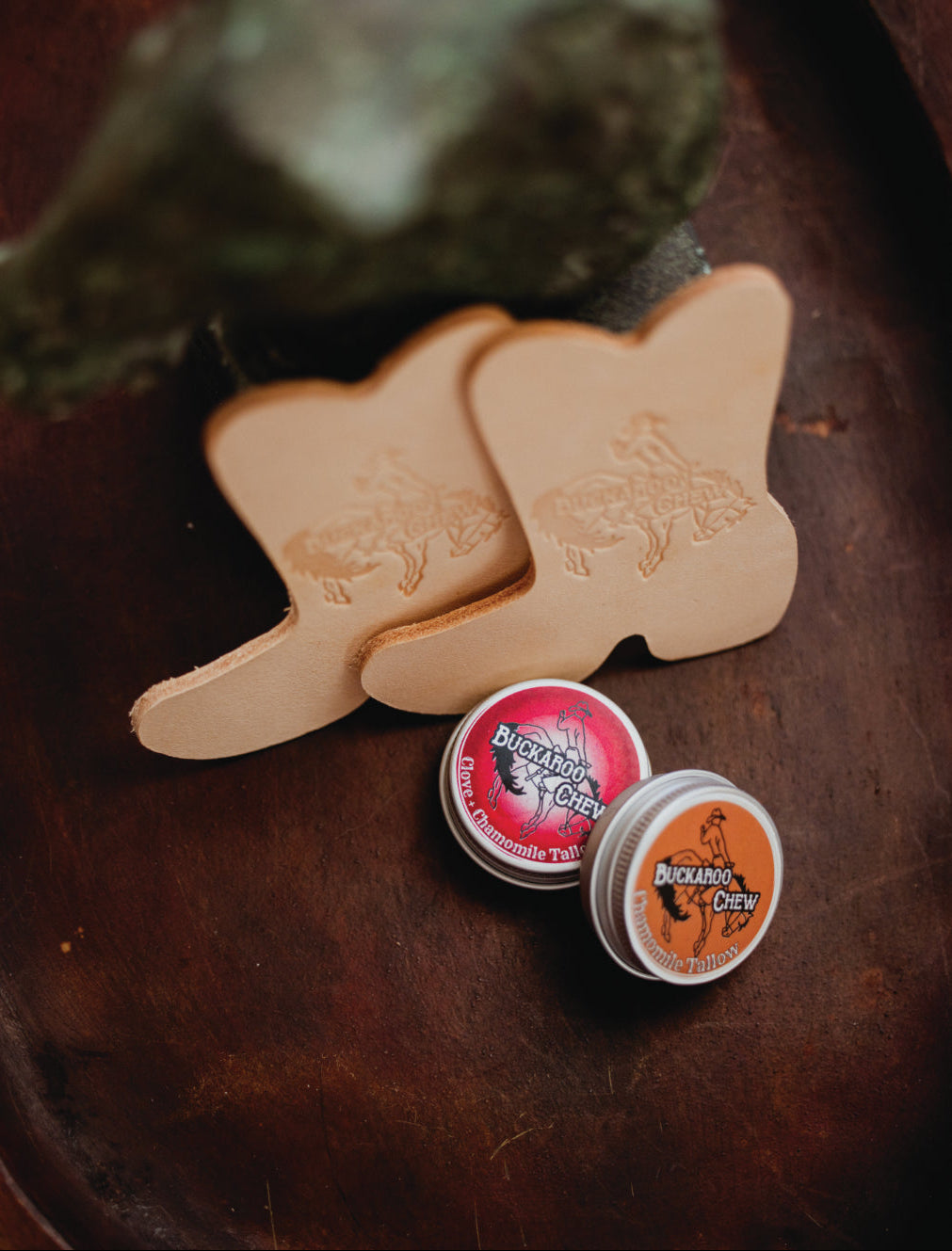 Two light tan leather boot-shaped pieces with embossed western designs, displayed on a dark brown wooden surface alongside two small round tins labeled Buckanoa Cider.