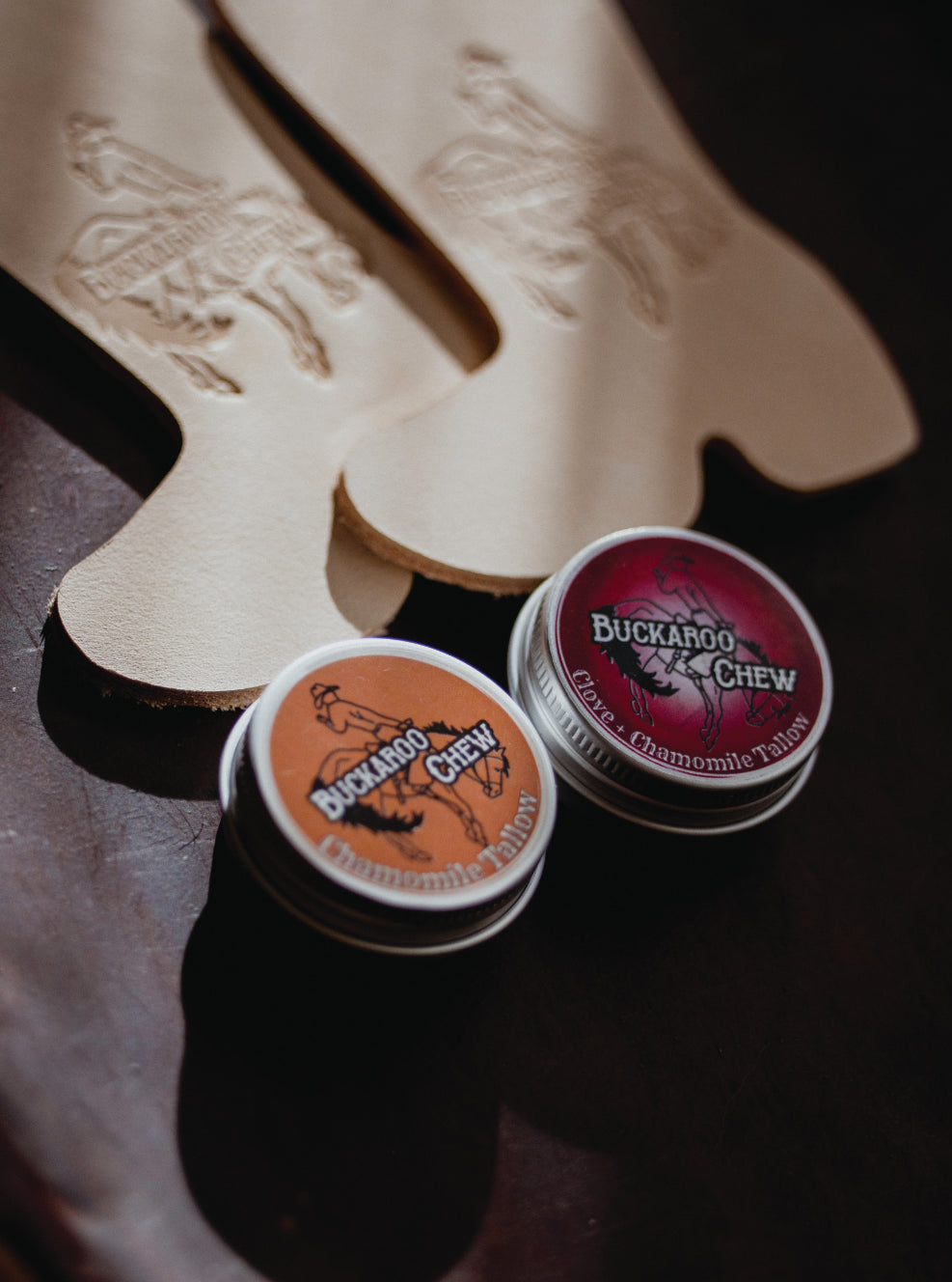 Two small round tins of Buckaroo Chew dog treats placed in front of a beige leather boot-shaped mat on a dark wooden surface. One tin has an orange label and the other has a red label, both reading Buckaroo Chew.