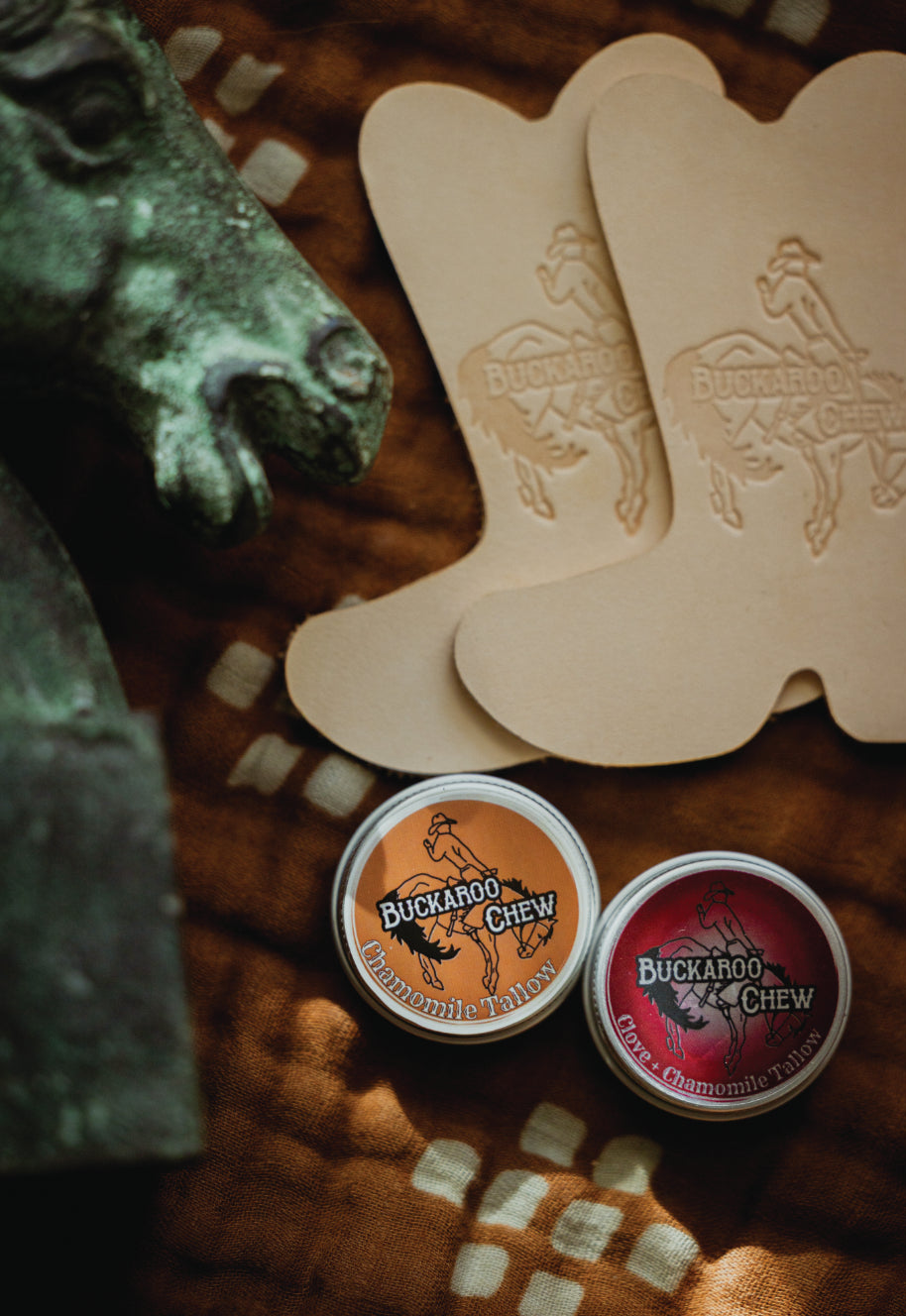 Two small round tins of Buckaroo Chew gummi tabs with orange and red labels, placed on a brown textured blanket beside two beige cowboy boot-shaped pieces embossed with the Buckaroo Chew logo. A dark green horse head sculpture is partially visible on the left.