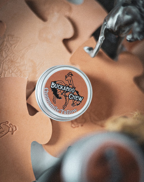 A small open tin of Chamomile Tallow with a label that has a beige background and features an illustration of a horse. The tin is placed on a surface with a leather object and a decorative horse figure in the background.